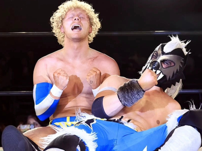 Two male wrestlers in a ring: a blond, curly-haired man screams with fists clenched while his partner, wearing a wolf-masks, grabs him in a hold.