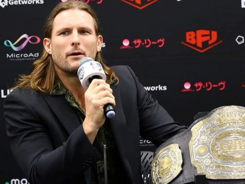 Man with shoulder-length hair in a blazer speaks into a microphone at a press conference, with a large championship belt visible in the foreground.