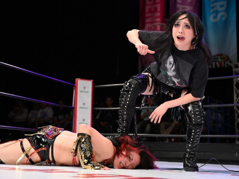 Wrestler in black with studded boots stands over a fallen opponent in a ring, gesturing toward her as the crowd watches.|A female wrestler in black gear looms over a downed rival on the mat, pointing accusatorily in a wrestling ring with ropes around them.| Standing wrestler in dark attire and makeup encroaches on a fallen opponent in a wrestling ring, pointing down as the referee or crowd looks on.| Female wrestlers clash: one person stands in black, while the other lies on the mat, the standing wrestler pointing in triumph or taunt inside the ring.