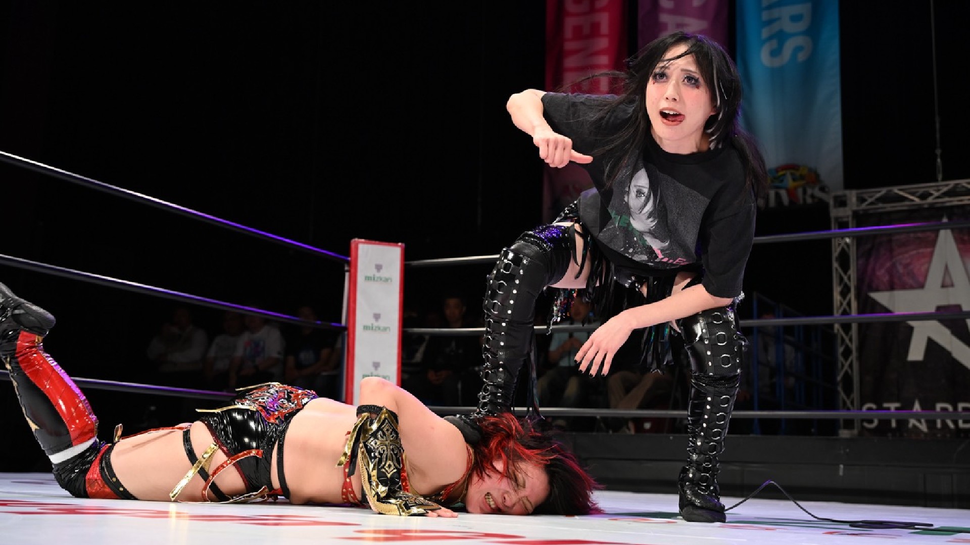 Wrestler in black with studded boots stands over a fallen opponent in a ring, gesturing toward her as the crowd watches.|A female wrestler in black gear looms over a downed rival on the mat, pointing accusatorily in a wrestling ring with ropes around them.| Standing wrestler in dark attire and makeup encroaches on a fallen opponent in a wrestling ring, pointing down as the referee or crowd looks on.| Female wrestlers clash: one person stands in black, while the other lies on the mat, the standing wrestler pointing in triumph or taunt inside the ring.