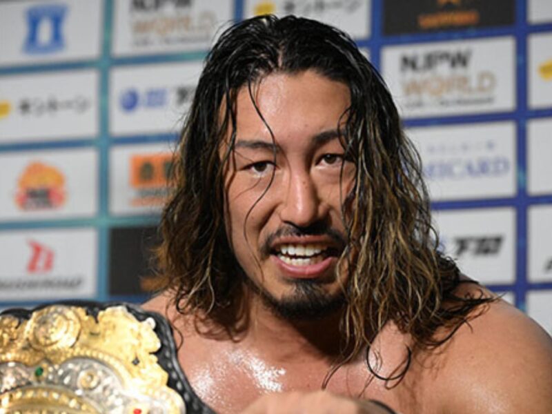 Wrestler with long hair smiling, holding a gold championship belt over his shoulder in front of sponsor boards.