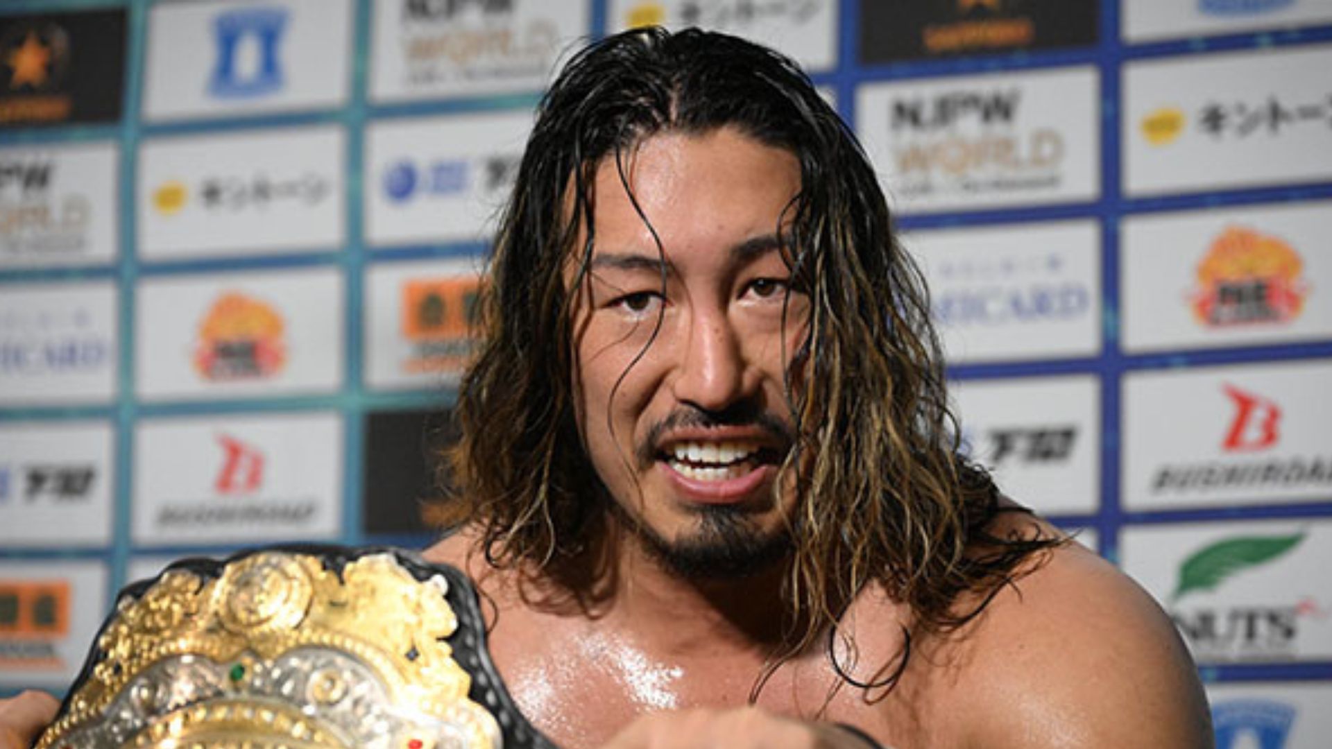 Wrestler with long hair smiling, holding a gold championship belt over his shoulder in front of sponsor boards.
