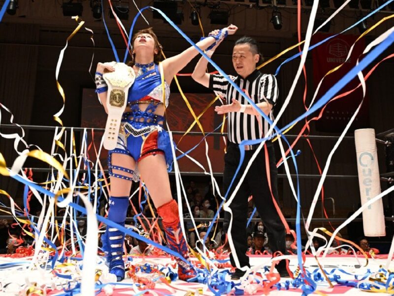 Wrestler in blue gear holds a championship belt as the referee raises her arm amid a shower of colorful streamers in the ring.