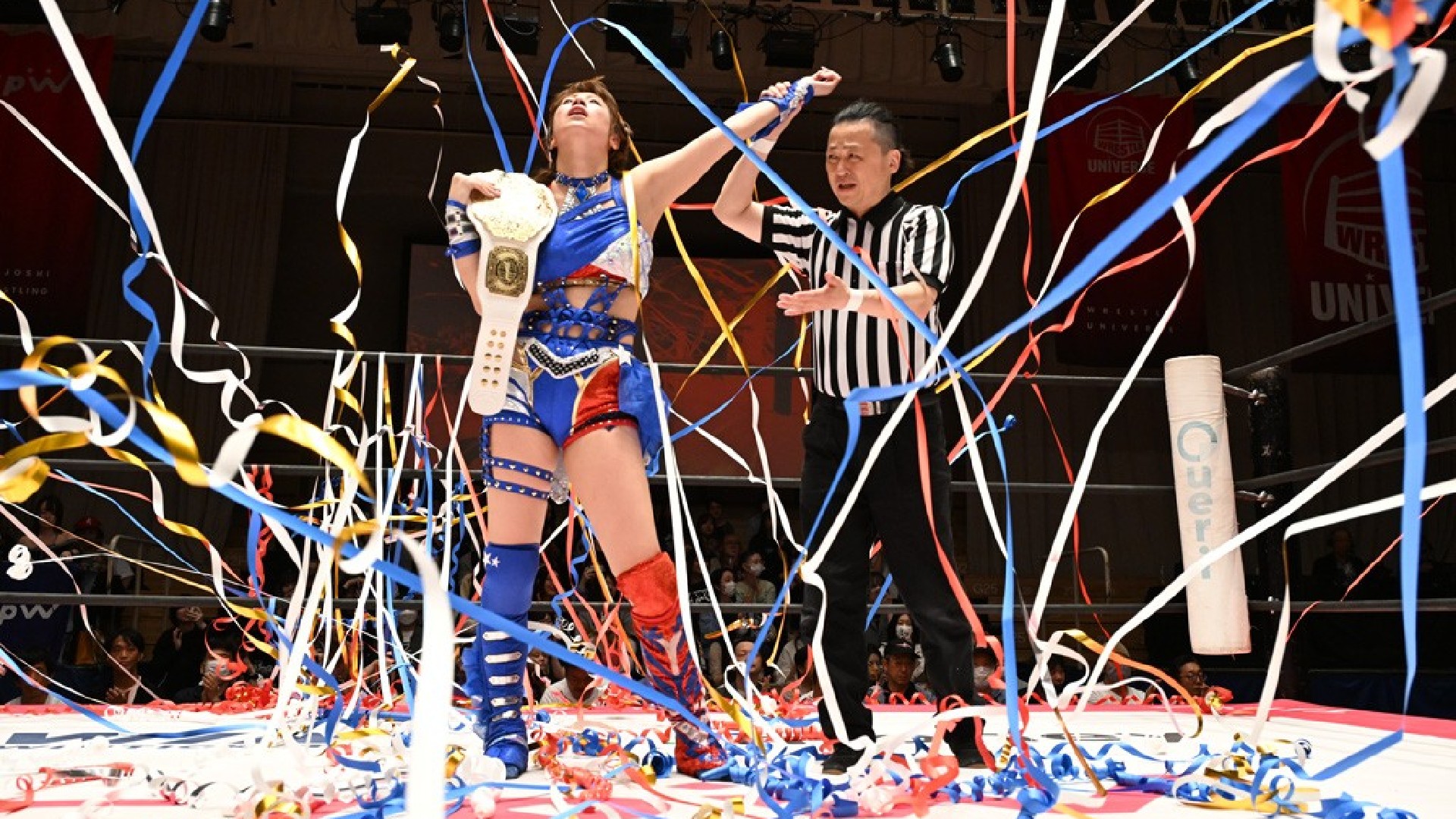 Wrestler in blue gear holds a championship belt as the referee raises her arm amid a shower of colorful streamers in the ring.