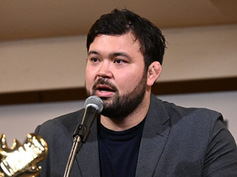Man with dark hair and beard wearing a gray blazer speaks into a microphone at a panel, with a gold trophy visible in the foreground.