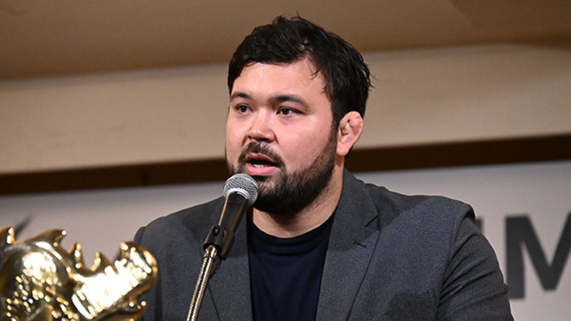 Man with dark hair and beard wearing a gray blazer speaks into a microphone at a panel, with a gold trophy visible in the foreground.