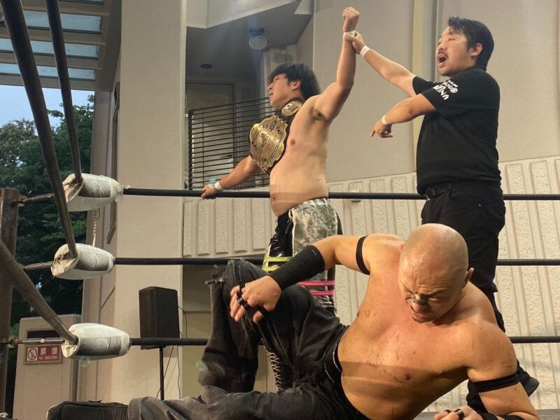Wrestler with championship belt raises arm in victory as teammate cheers inside the ring. Another wrestler grimaces in the foreground.