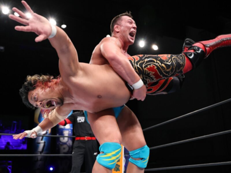 Two wrestlers collide mid-match as one lifts the other overhead in the ring, faces intense with effort.