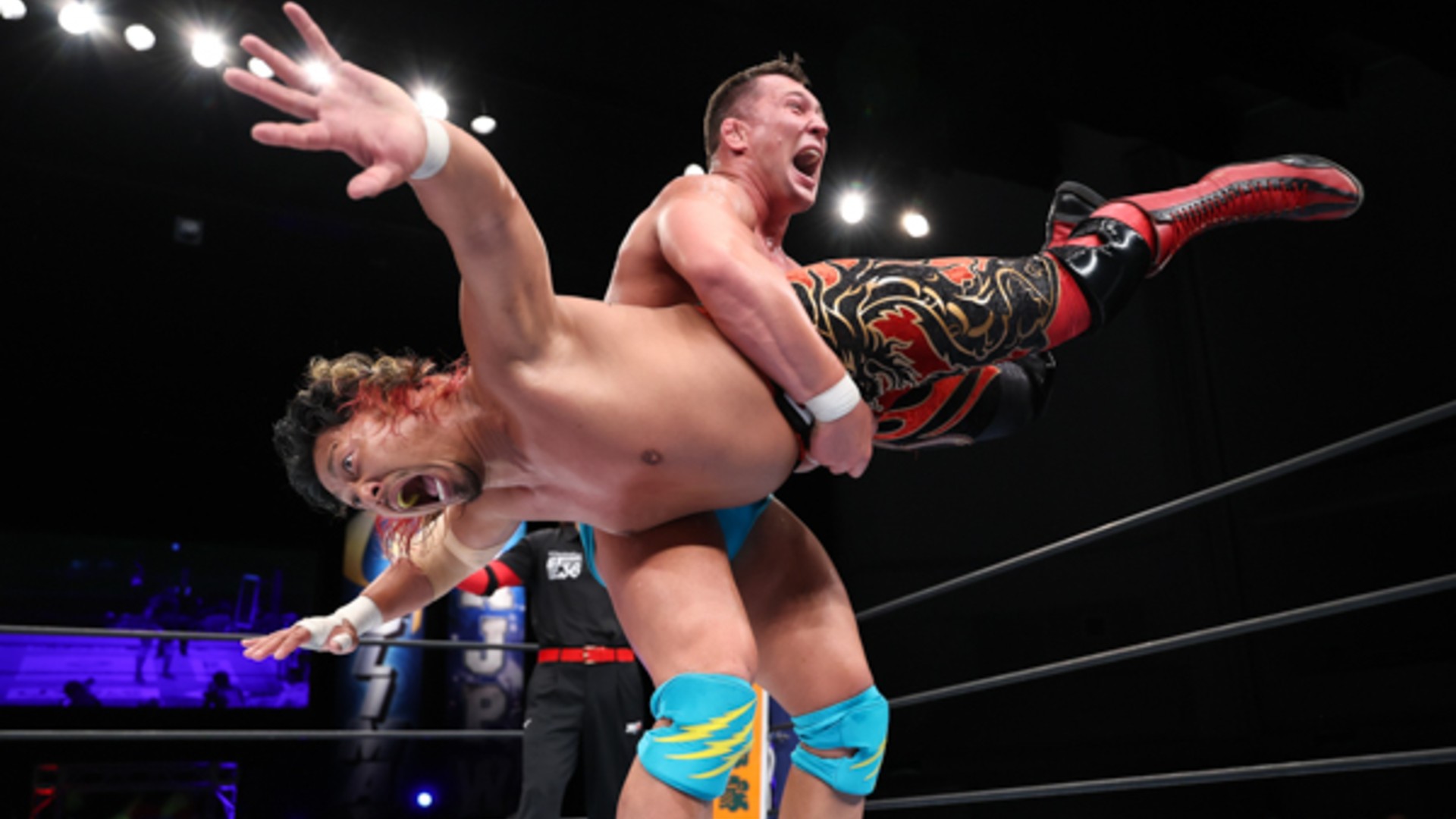 Two wrestlers collide mid-match as one lifts the other overhead in the ring, faces intense with effort.