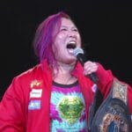 Female wrestler with pink-dyed hair wearing a red jacket, shouting into a microphone while holding a large championship belt.