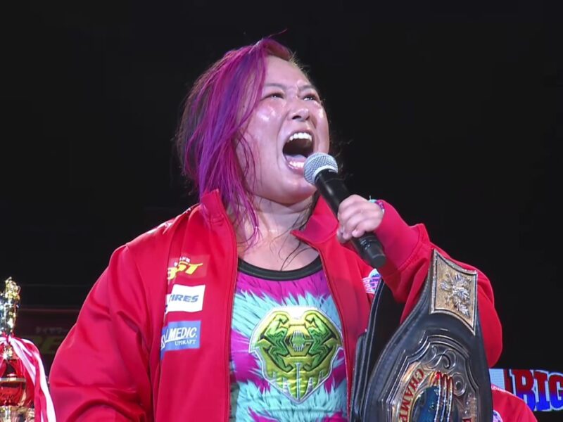Female wrestler with pink-dyed hair wearing a red jacket, shouting into a microphone while holding a large championship belt.