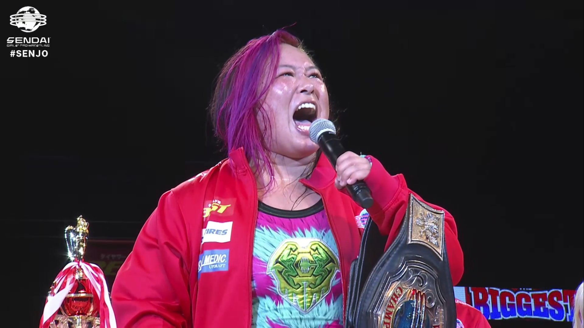Female wrestler with pink-dyed hair wearing a red jacket, shouting into a microphone while holding a large championship belt.