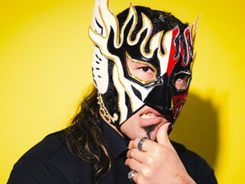 Person wearing a black shirt and a decorative black, white, and red lucha libre mask against a bright yellow background
