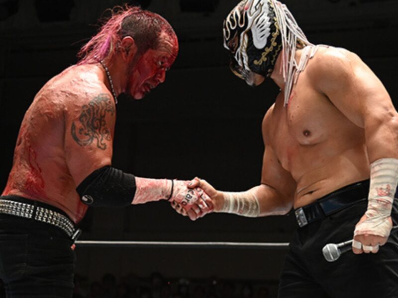 Wrestlers shake hands in a ring after a match; one has red body paint and tattoos, the other wears a decorative mask and holds a microphone.