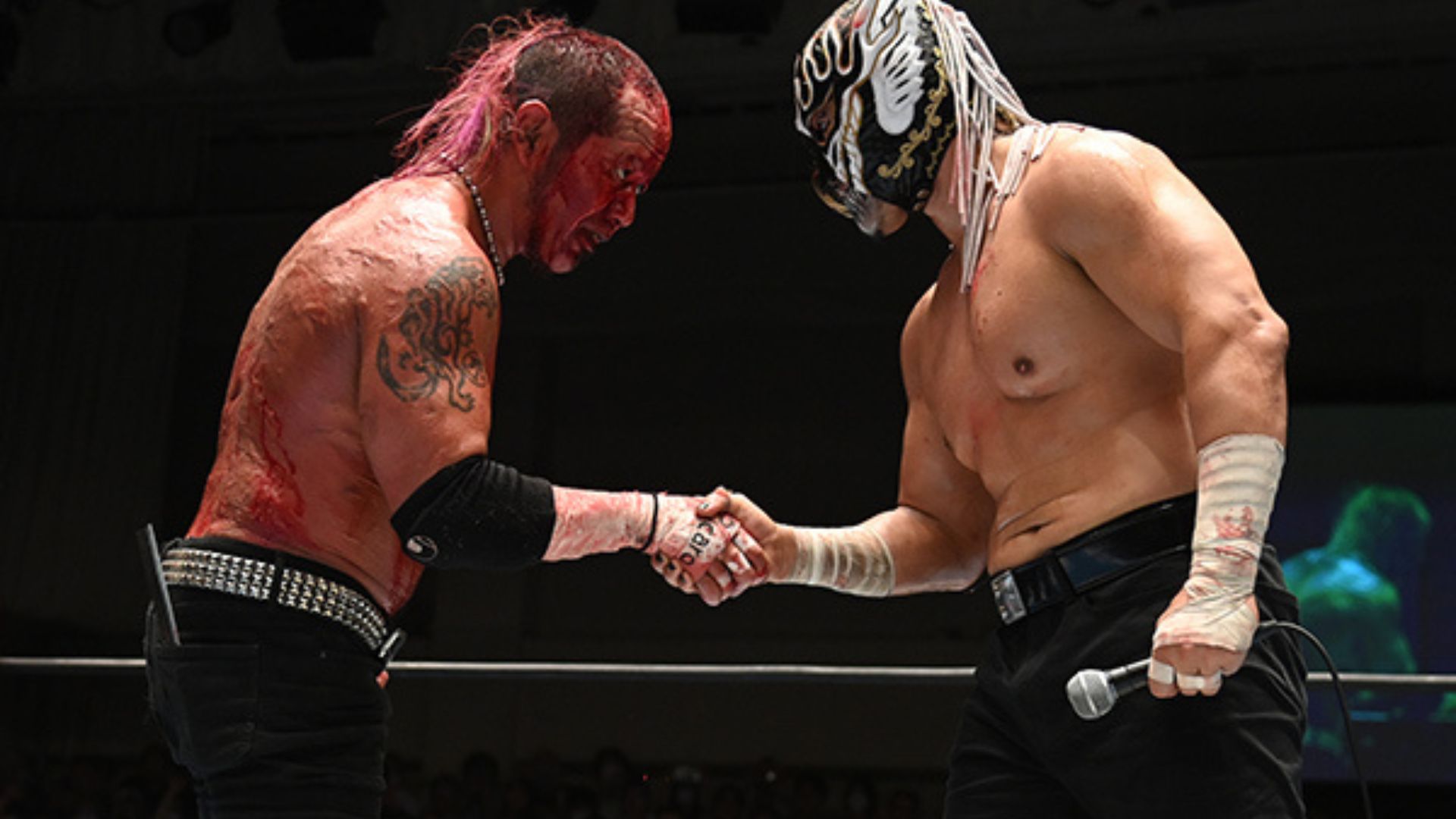 Wrestlers shake hands in a ring after a match; one has red body paint and tattoos, the other wears a decorative mask and holds a microphone.