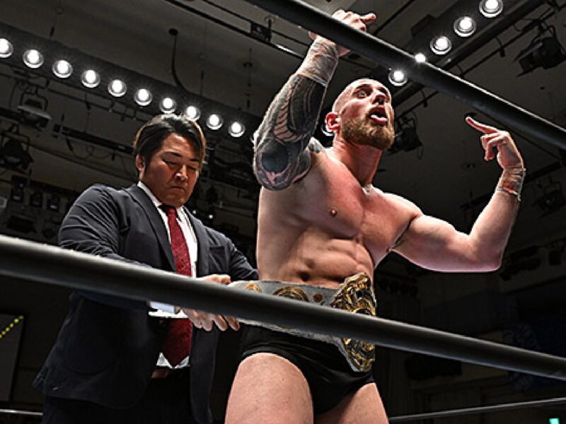 Wrestler with a championship belt stands in the ring, flexing at the camera while a suited official leans on the ropes nearby.