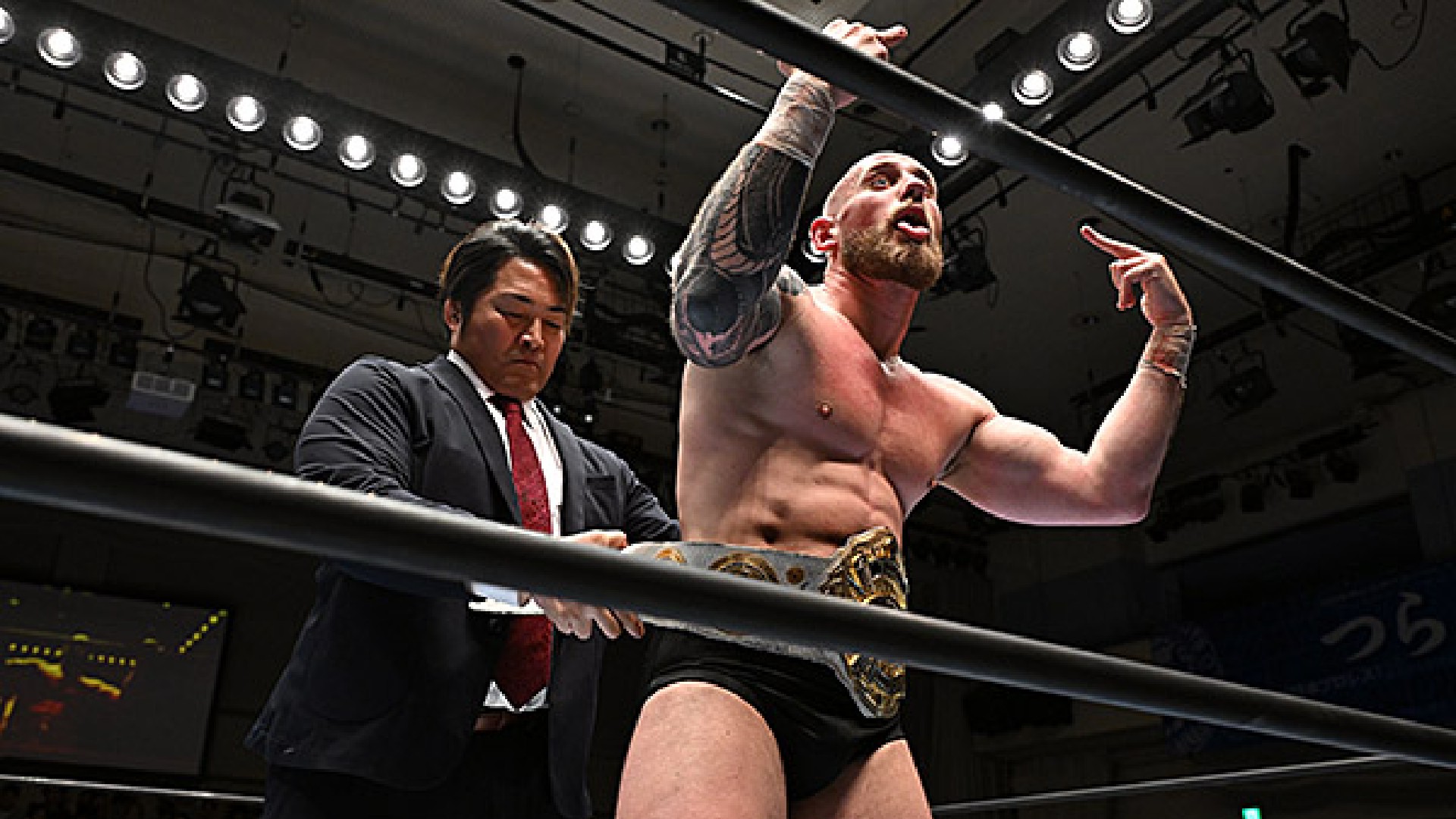 Wrestler with a championship belt stands in the ring, flexing at the camera while a suited official leans on the ropes nearby.