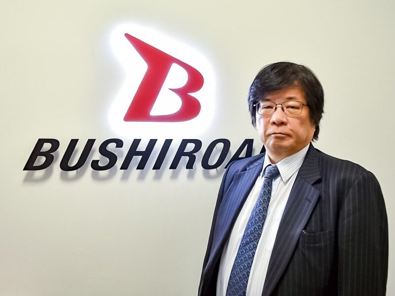 Man in a dark pinstripe suit and glasses stands beside a Bushiroad logo on a light wall.