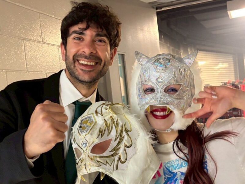 Smiling man in a suit and two people in silver masquerade masks pose for a selfie backstage.