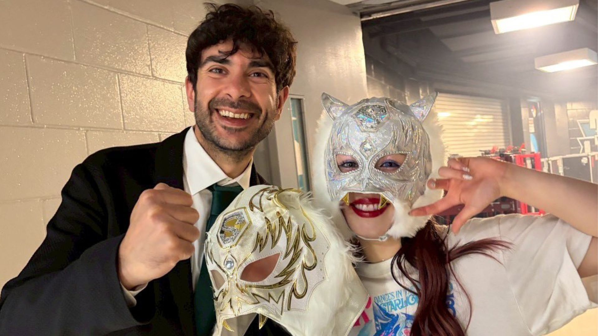 Smiling man in a suit and two people in silver masquerade masks pose for a selfie backstage.