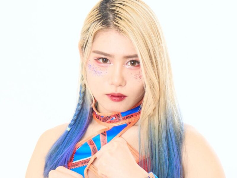Portrait of a young woman with long blonde hair and blue‑tinted ends, wearing a blue/orange caution‑tape scarf and glitter freckles on her cheeks.
