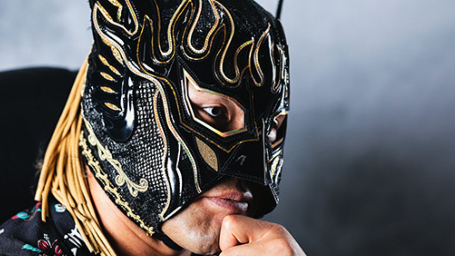 Close-up of a person wearing a black and gold ornate luchador mask, hand resting on chin.