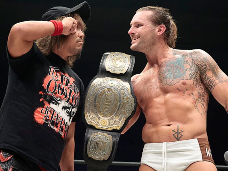 Two professional wrestlers in a ring celebrate; one wears a large championship belt draped over his shoulder while the other faces him
