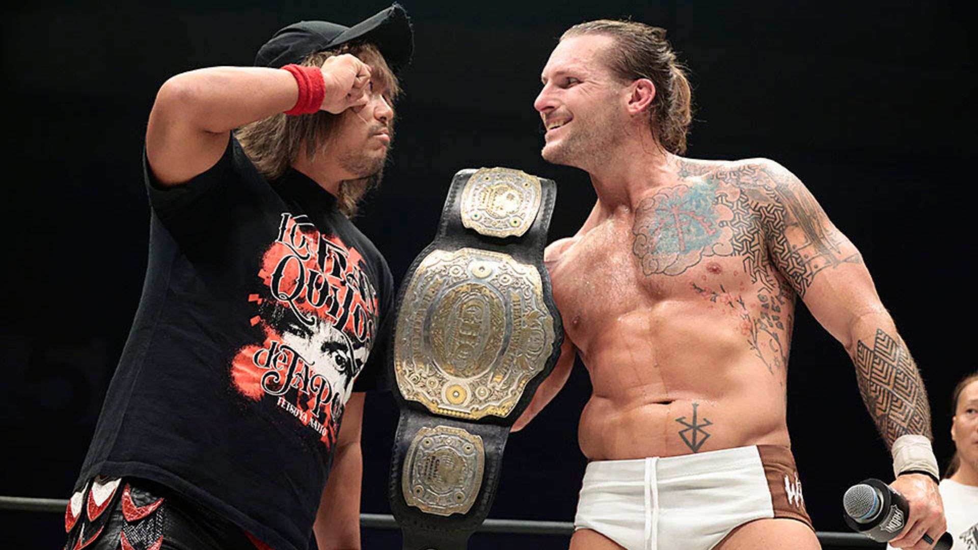 Two professional wrestlers in a ring celebrate; one wears a large championship belt draped over his shoulder while the other faces him