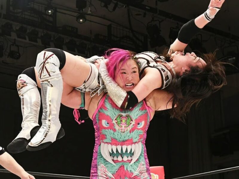 Wrestler with pink hair lifts her opponent overhead inside a ring while a referee watches to the left.