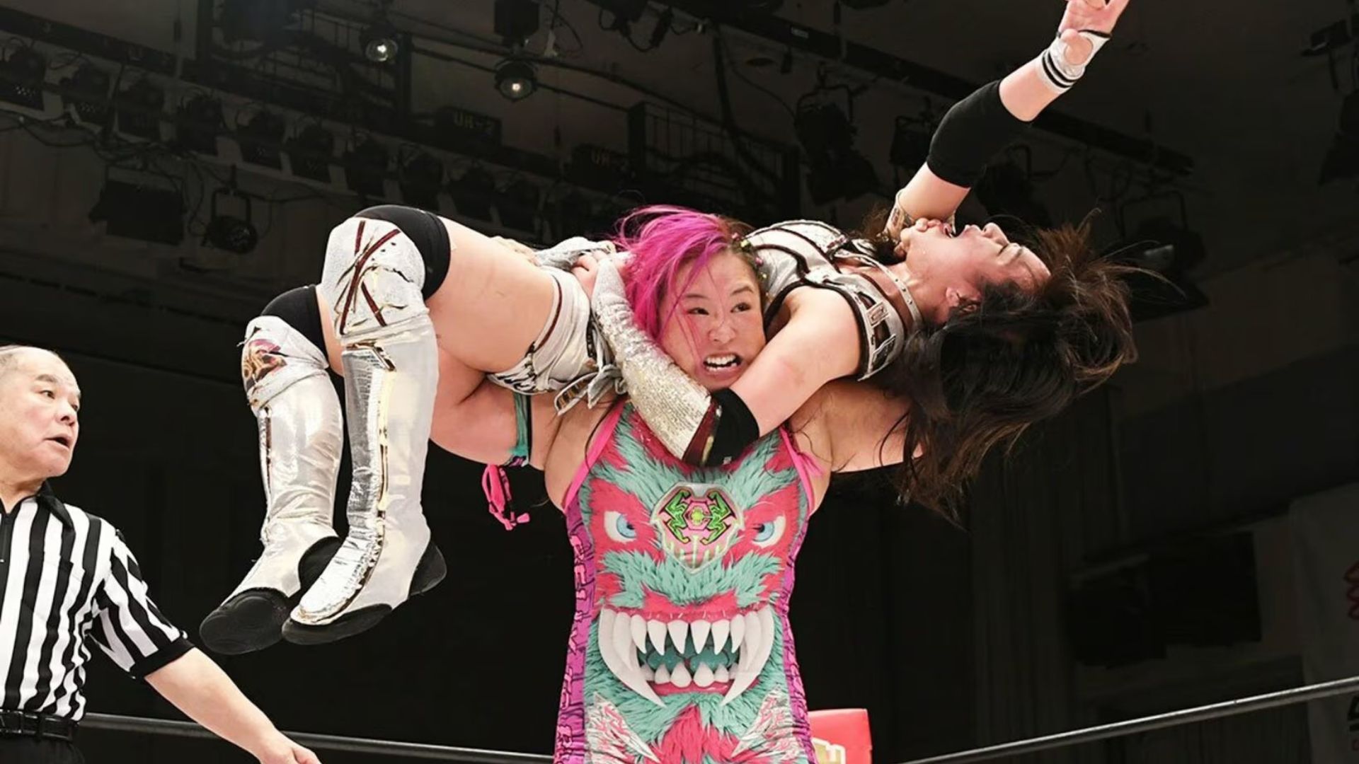 Wrestler with pink hair lifts her opponent overhead inside a ring while a referee watches to the left.