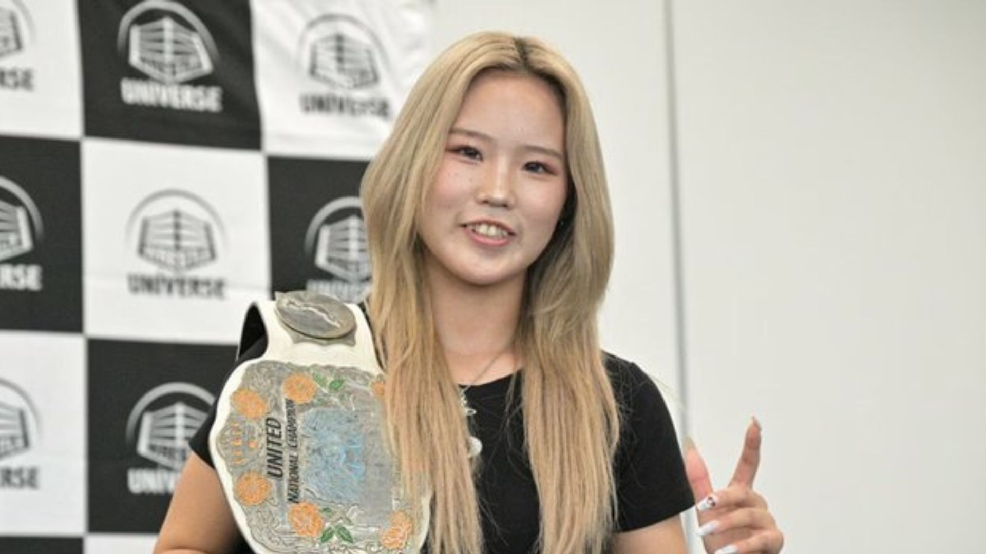 Smiling young woman wearing a decorative championship belt, posing in front of a checkered backdrop with logos.