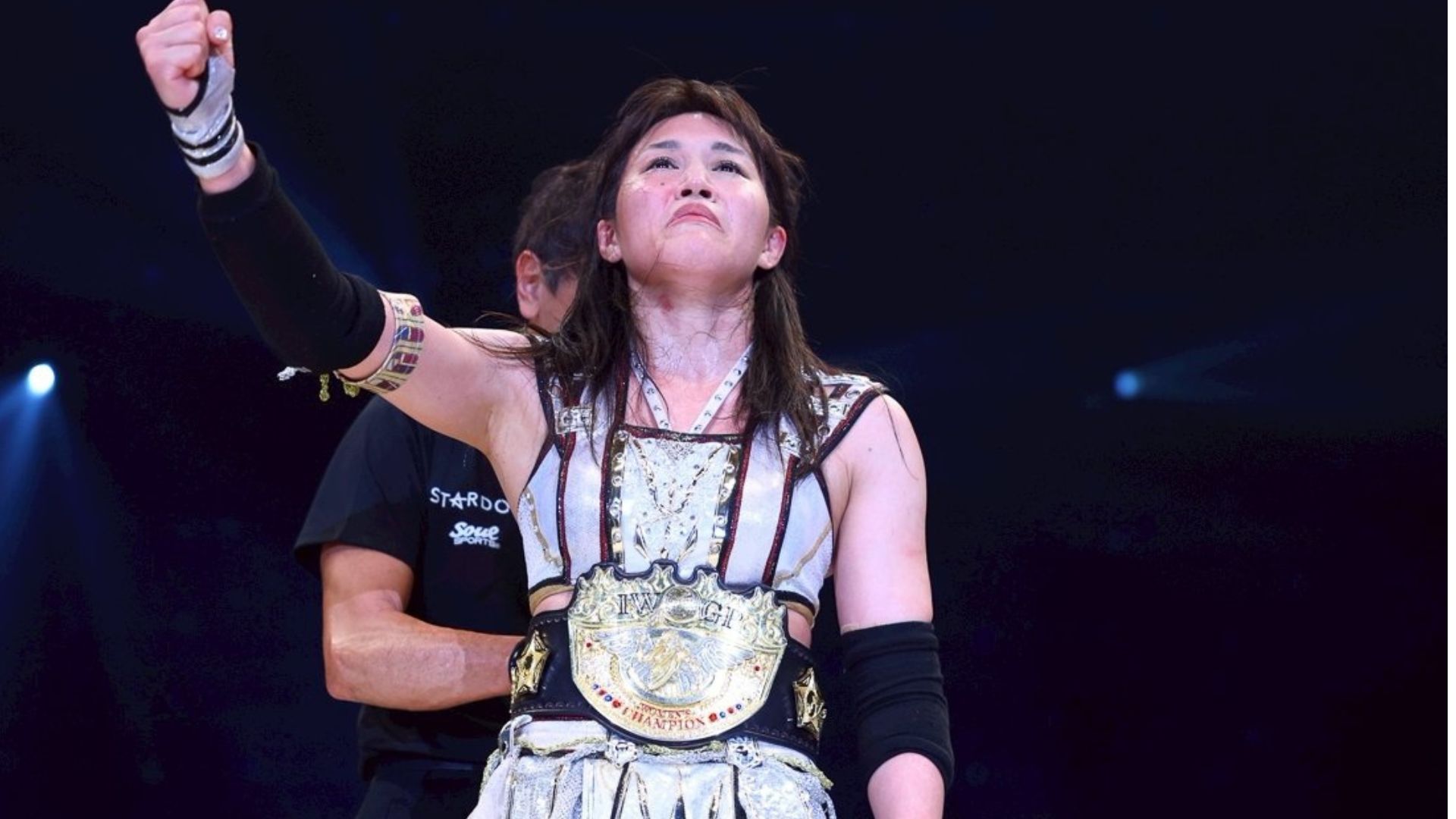 Female wrestler on stage, raising her right fist in victory while wearing a large championship belt.