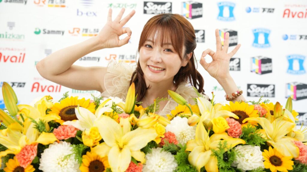 Smiling woman behind a large bouquet of yellow flowers, making OK signs with both hands in front of a sponsor backdrop.