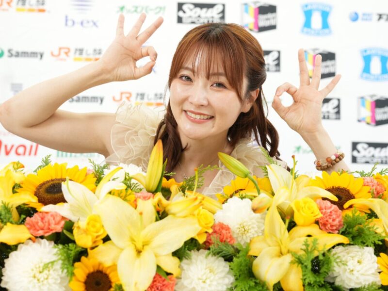 Smiling woman behind a large bouquet of yellow flowers, making OK signs with both hands in front of a sponsor backdrop.
