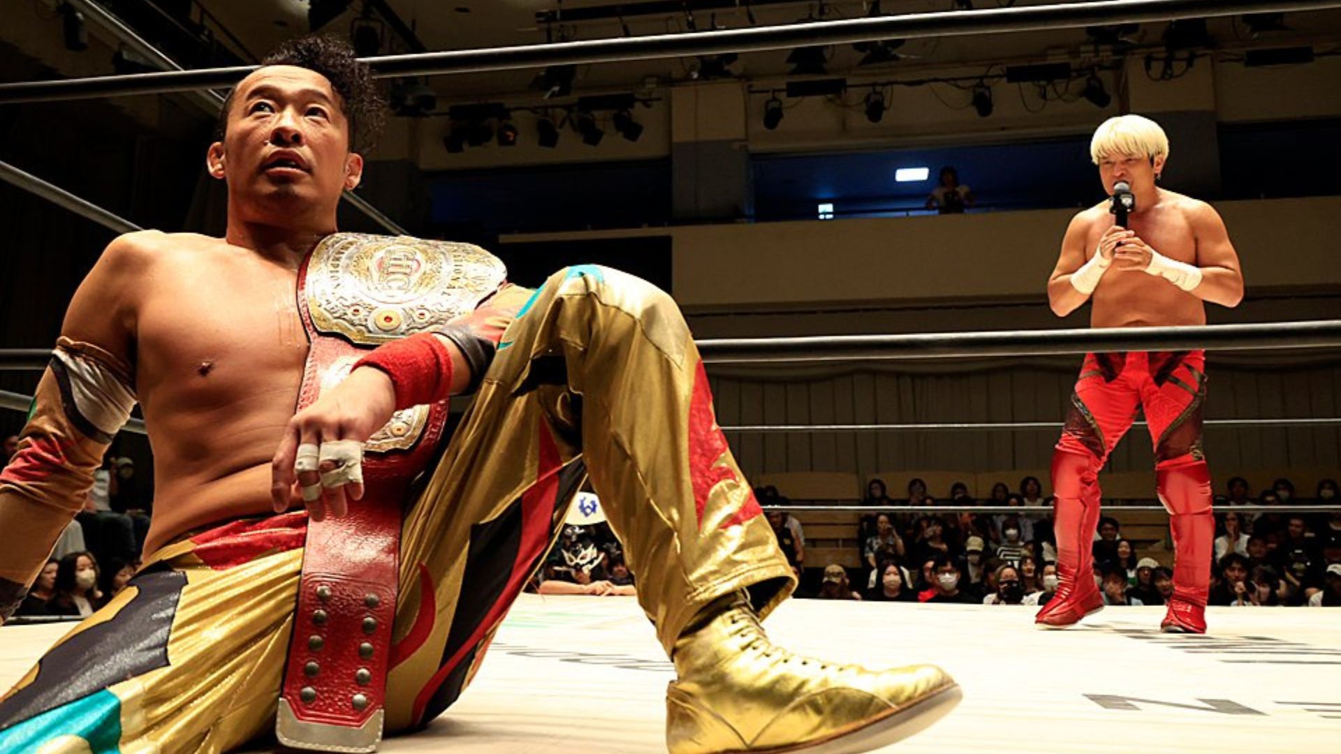 Wrestlers in a ring during a match; one man sits on the mat with a gold championship belt, while another stands with a microphone as spectators watch.