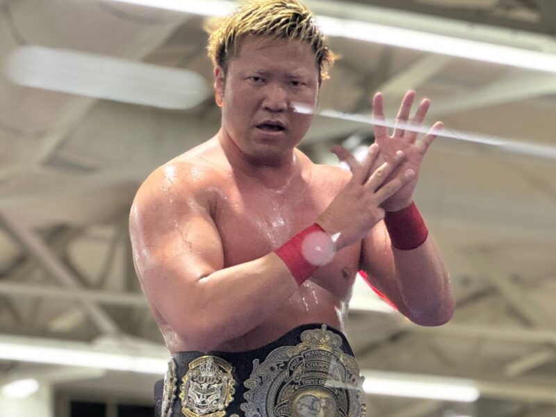 Muscular wrestler wearing a large championship belt, standing in a arena with hands pressed together in front of his chest.