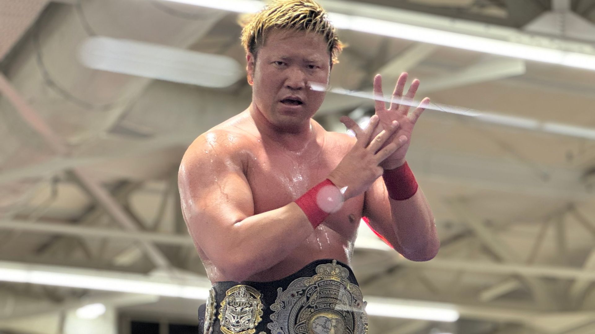 Muscular wrestler wearing a large championship belt, standing in a arena with hands pressed together in front of his chest.