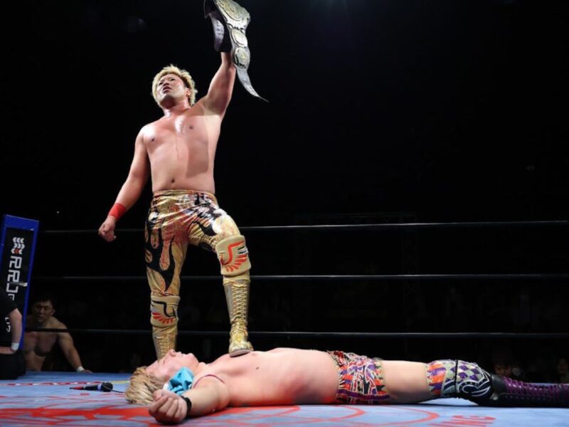 Wrestler stands over a fallen opponent in a ring, raising a championship belt in victory as spectators watch in the background.