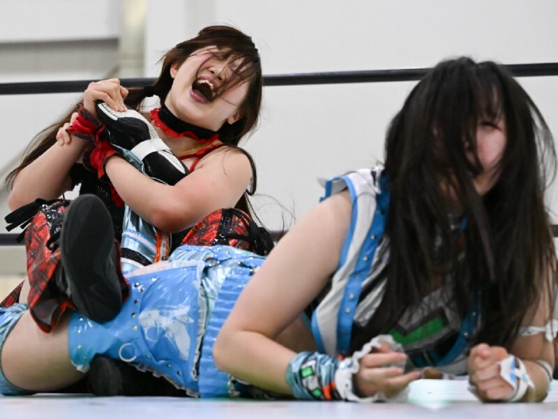 Two female wrestlers clash in a ring, one laughing wildly as she gets tangled in colorful costumes while the other is on the mat.