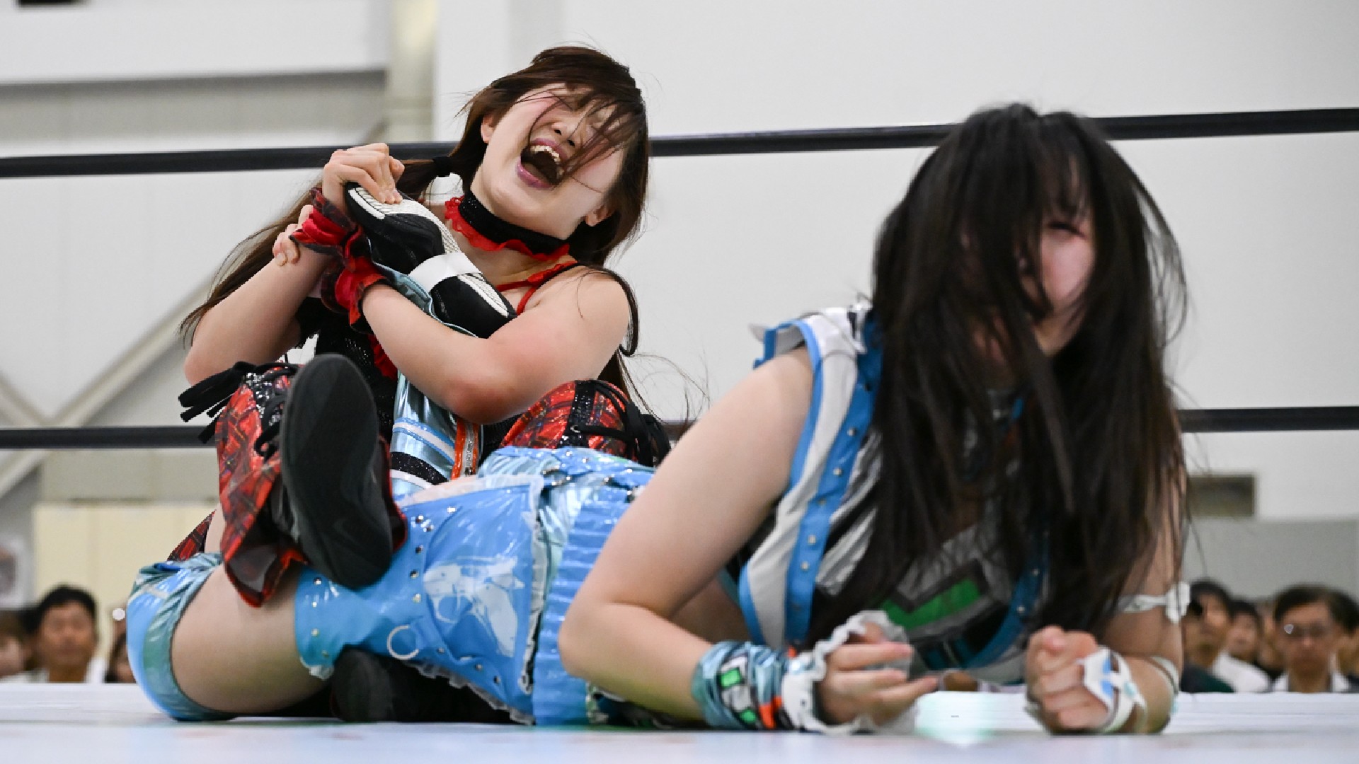 Two female wrestlers clash in a ring, one laughing wildly as she gets tangled in colorful costumes while the other is on the mat.