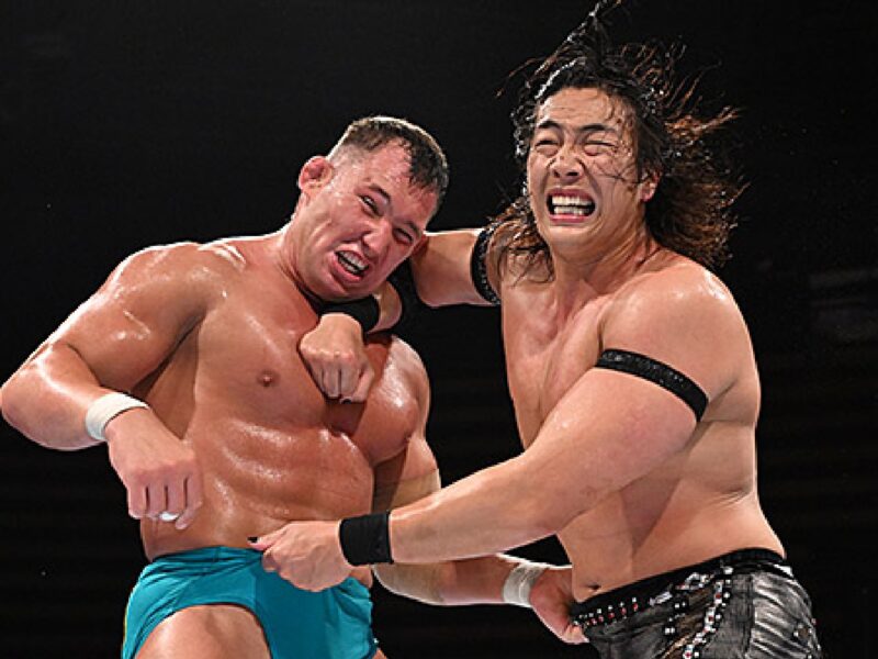 Two wrestlers exchange punches in a ring, one wearing turquoise trunks and the other in black-patterned shorts, both grimacing from impact.