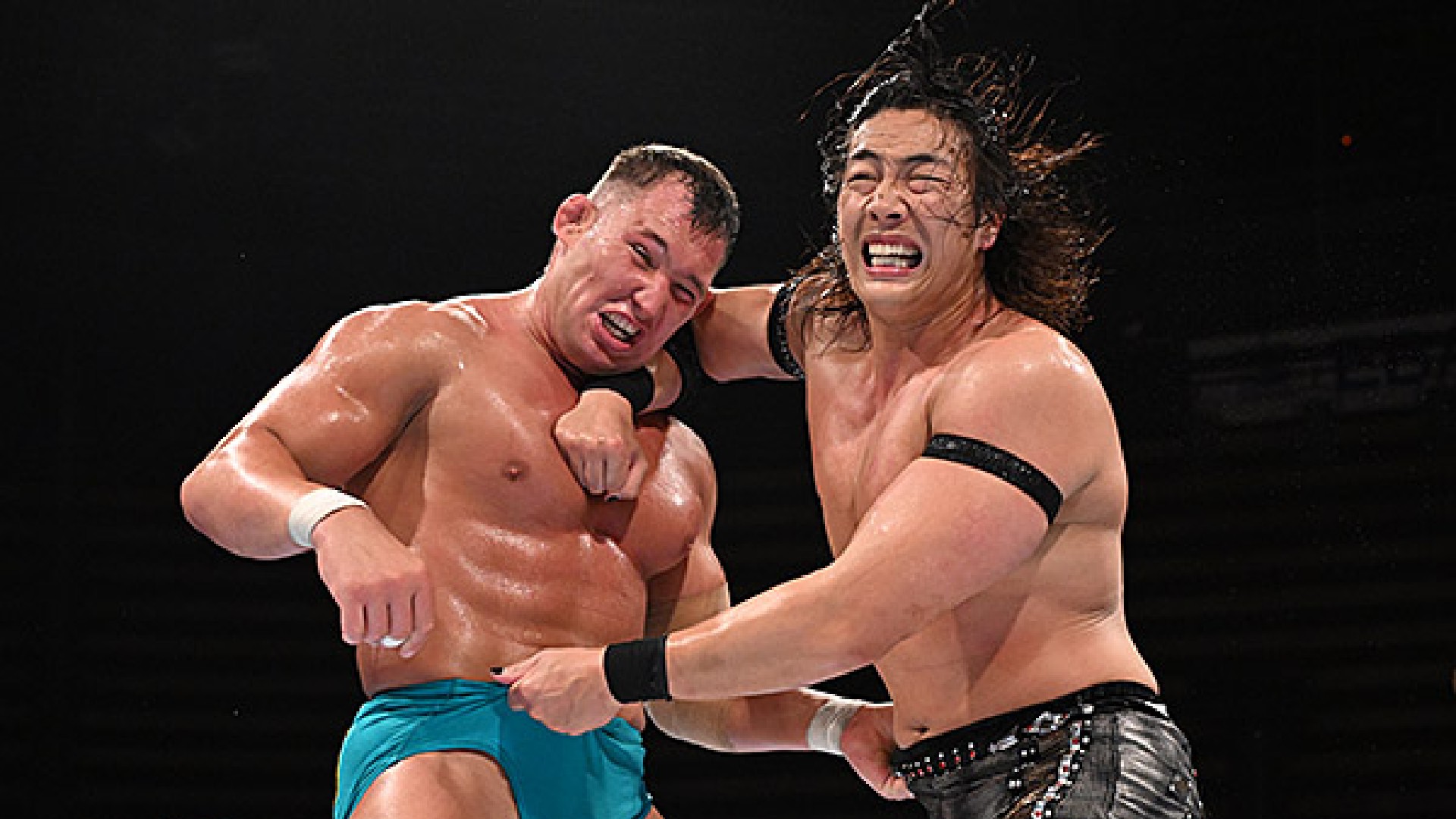 Two wrestlers exchange punches in a ring, one wearing turquoise trunks and the other in black-patterned shorts, both grimacing from impact.