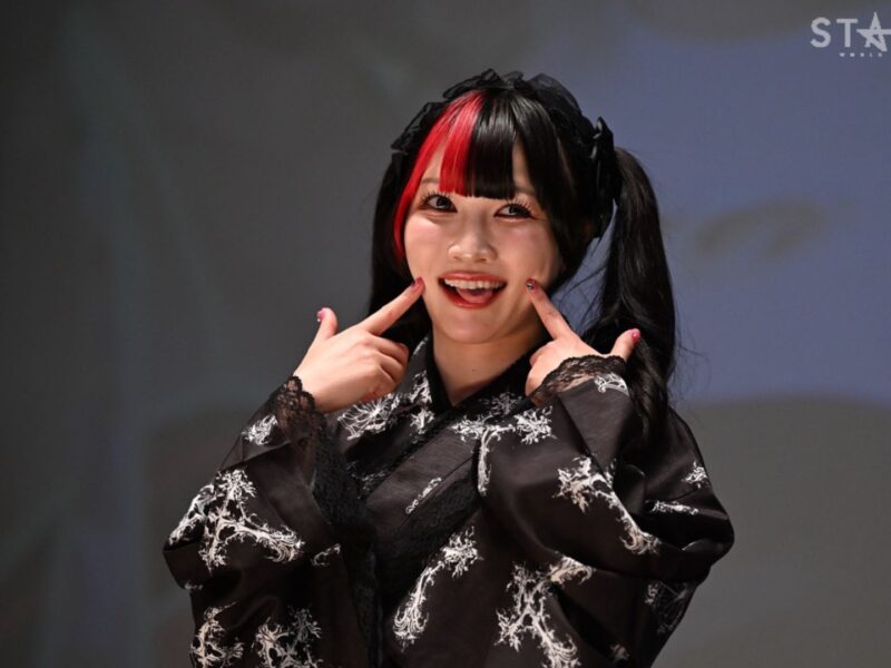 Young woman in a black kimono with white patterns, red and black hair, posing with fingers on her cheeks; Stardom logo in the background.