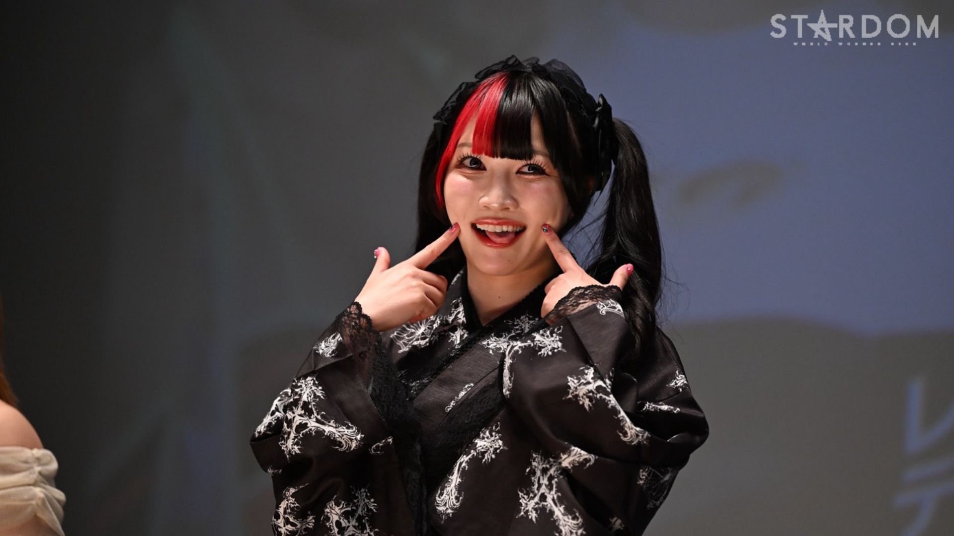 Young woman in a black kimono with white patterns, red and black hair, posing with fingers on her cheeks; Stardom logo in the background.