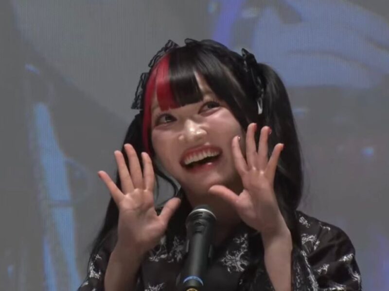 Smiling young woman with black-and-red hair at a microphone on stage, hands raised in a cheerful pose.