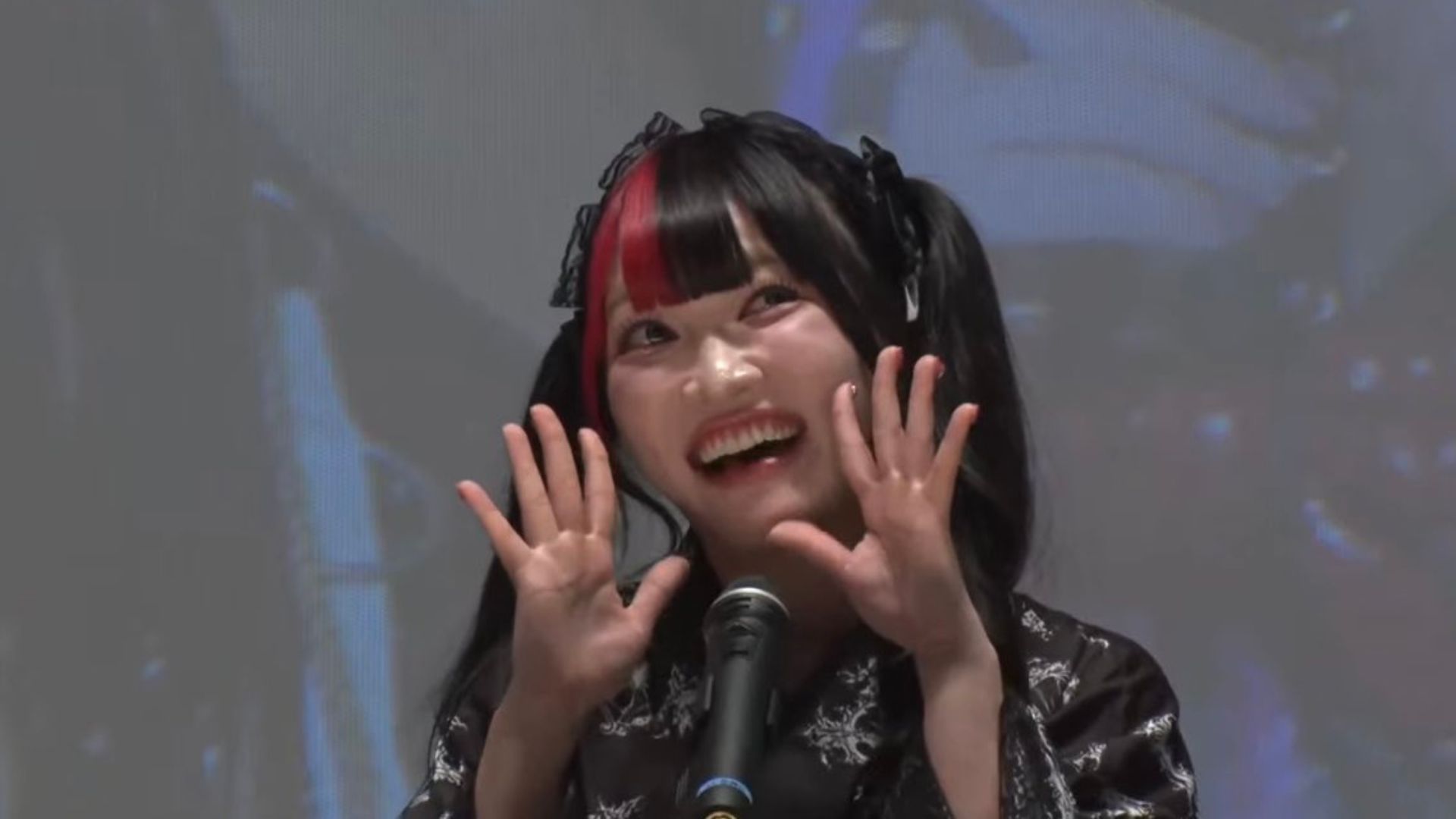 Smiling young woman with black-and-red hair at a microphone on stage, hands raised in a cheerful pose.