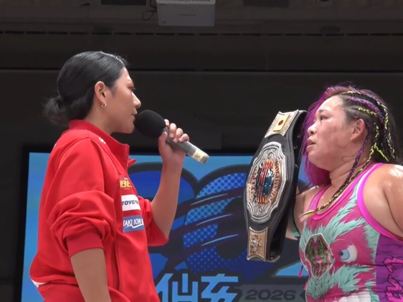 Female wrestler in a pink-green outfit, wearing a championship belt, speaks into a microphone during an interview.