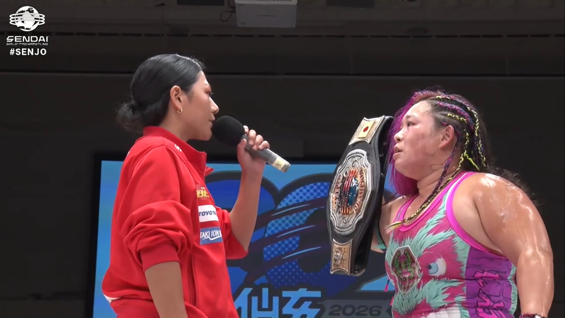 Female wrestler in a pink-green outfit, wearing a championship belt, speaks into a microphone during an interview.