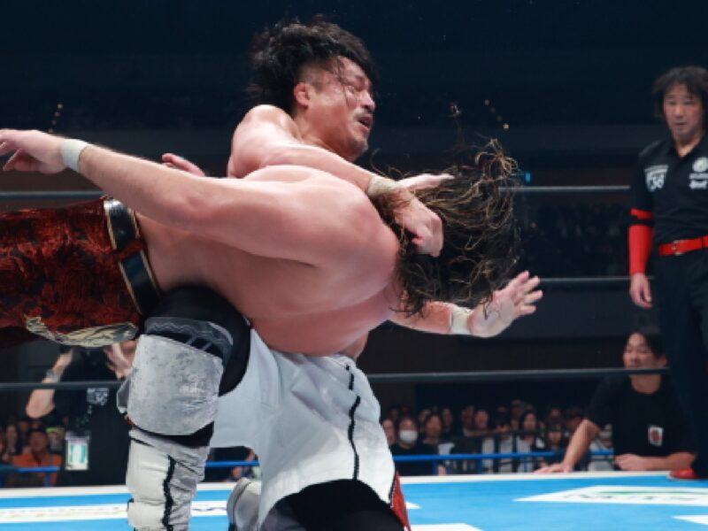 Two wrestlers grapple in the ring while a referee and audience watch from the side.