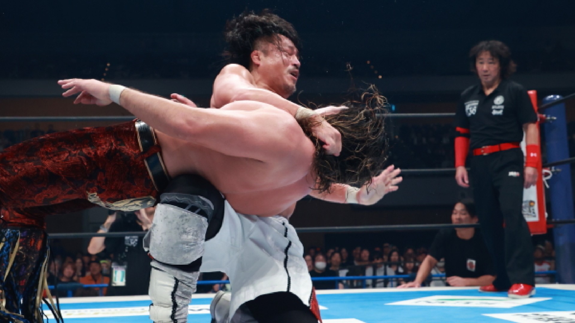 Two wrestlers grapple in the ring while a referee and audience watch from the side.